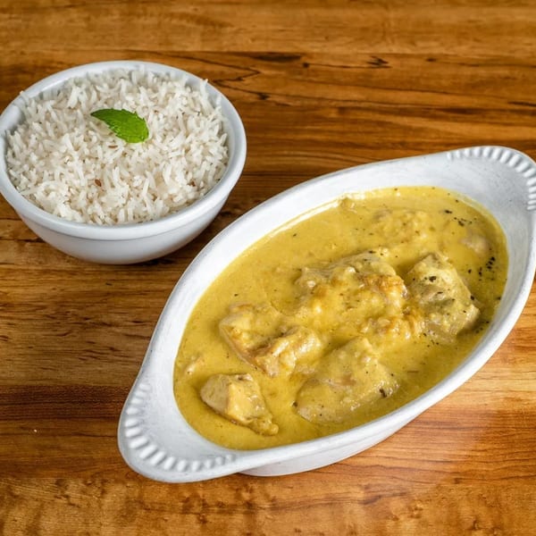 Creamy yellow chicken curry in a white dish next to a bowl of white rice with a mint leaf.