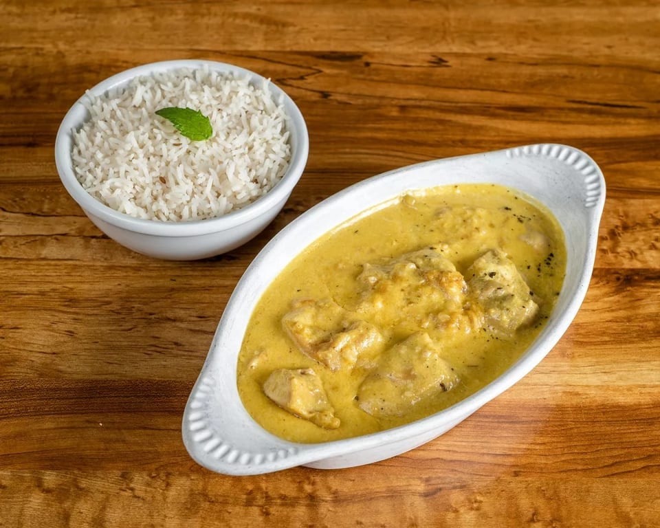 Creamy yellow chicken curry in a white dish next to a bowl of white rice with a mint leaf.