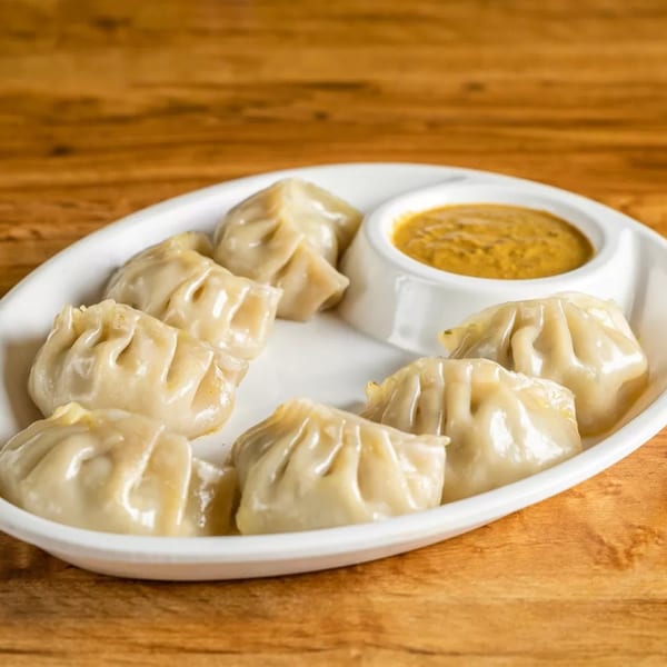 A white plate of eight steamed dumplings with a side of yellow dipping sauce on a wooden table.
