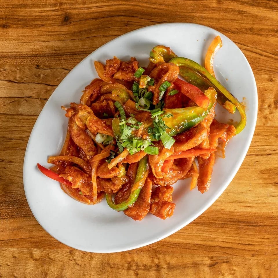 Chili chicken with bell peppers and onions, garnished with green onions, on a white oval plate.