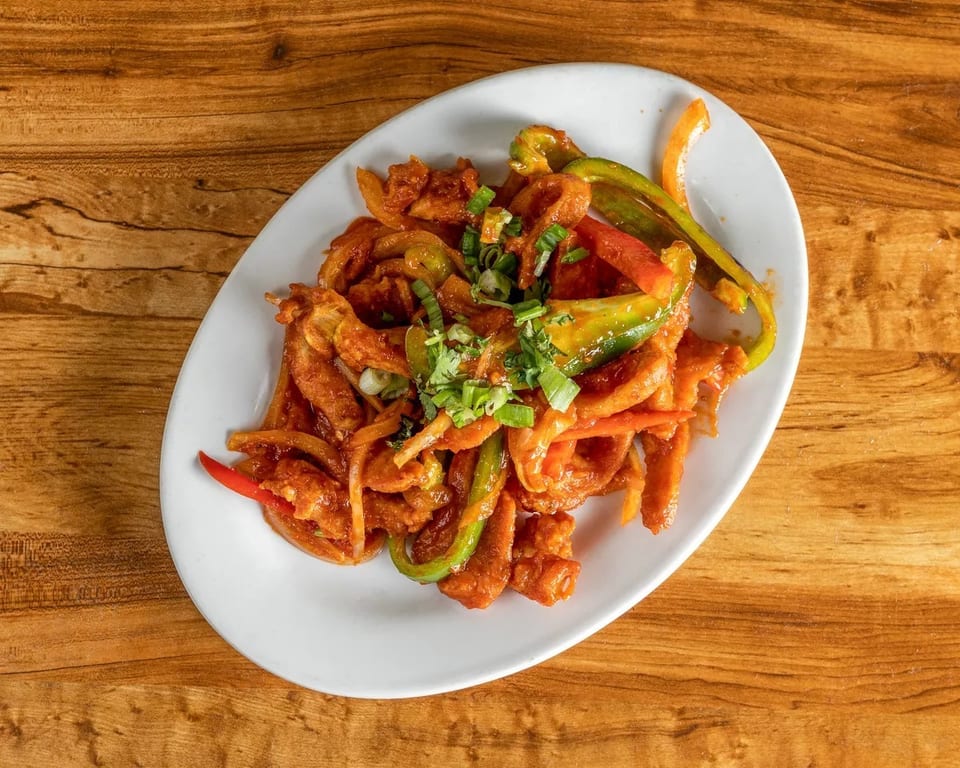 Chili chicken with bell peppers and onions, garnished with green onions, on a white oval plate.