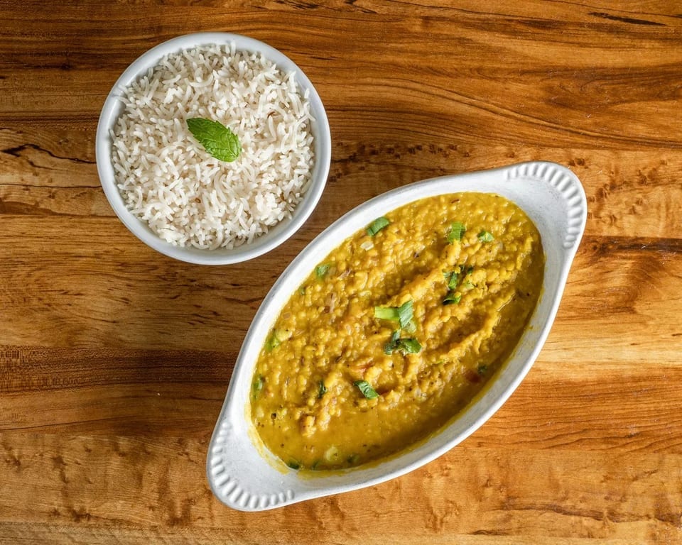 A bowl of white rice with a mint leaf and a dish of yellow lentil curry on a wooden table.