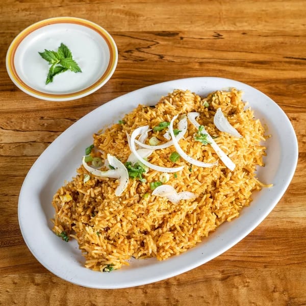 Golden biryani rice with sliced onions and green herbs, served with a side of raita.