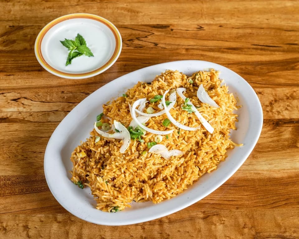 Golden biryani rice with sliced onions and green herbs, served with a side of raita.